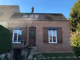 Maison en brique à volets bruns, un homme debout sur le faîtage nettoie le versant à la lance