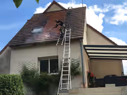 Intervenant sur une échelle contre le pignon, nettoyant les tuiles autour d’une fenêtre de toit